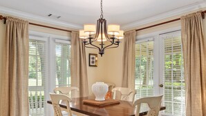 Comedor con mesa de madera, cuatro sillas, una lámpara de araña y ventanas grandes con cortinas.