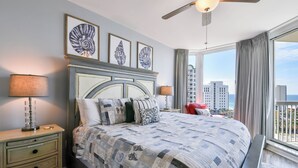 Ein Schlafzimmer mit einem großen Bett, einem Nachttisch, einem Deckenventilator und Blick auf die Stadt durch große Fenster.