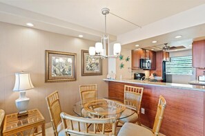 A dining area with a round glass table, wicker chairs, a chandelier, framed pictures on the wall, and a kitchen area with wooden cabinets and a refrigerator. - Oceanfront Oasis with Captivating Views: Royal Mauian 317 - Make Memories ! (Kihei, Maui)