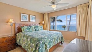 Un dormitorio con cama, ventilador de techo, ventana con vista al océano y mesita de noche.