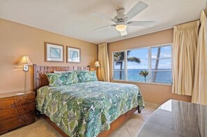 A bedroom with a bed, a ceiling fan, a window with a view of the ocean, and a nightstand. - Oceanfront Oasis with Captivating Views: Royal Mauian 317 - Make Memories ! (Kihei, Maui)