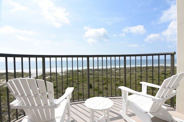 A balcony with two white Adirondack chairs and a small round table, overlooking a beach and ocean.