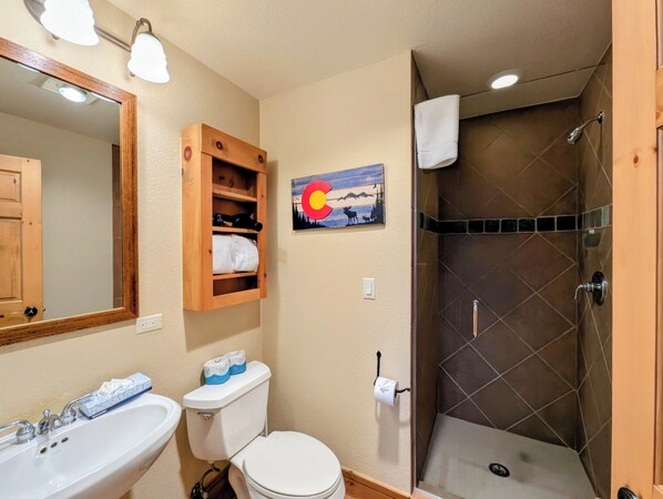 A bathroom with a sink, toilet, and shower. There is a wooden cabinet above the toilet and a painting on the wall. - Mountain Views + Pool | Shuttle to Slopes (Keystone)