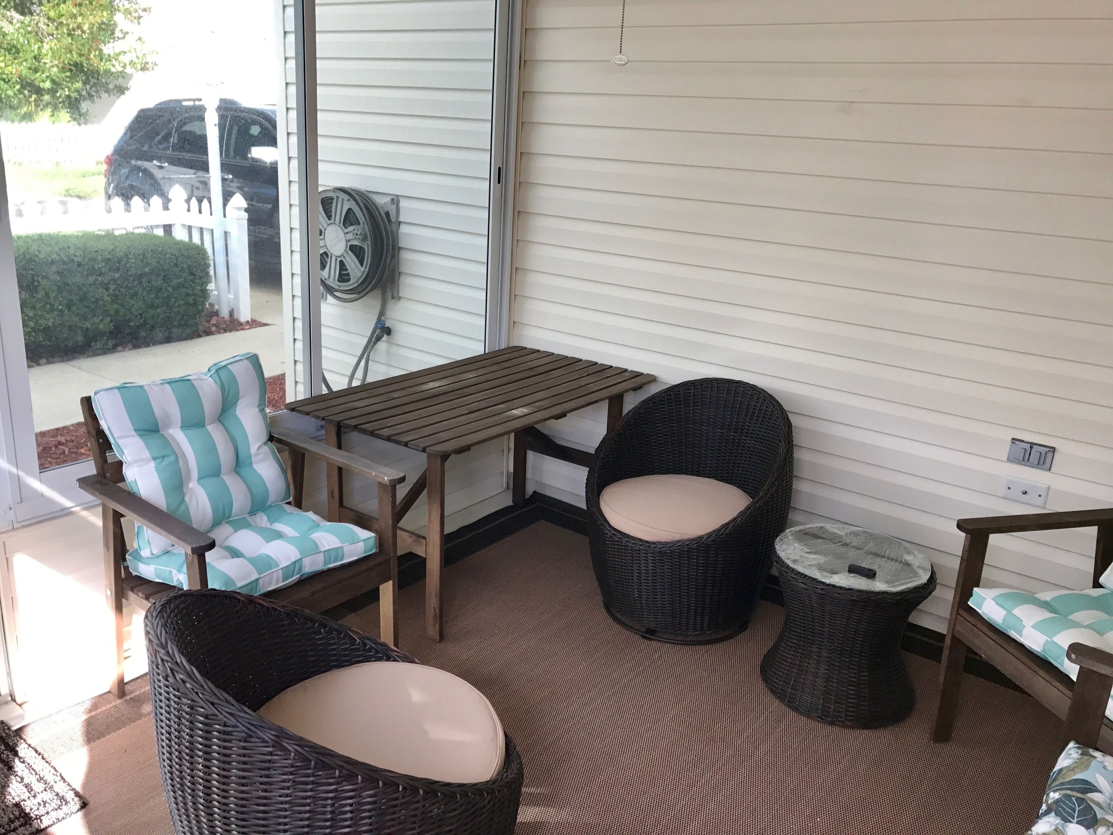 A covered patio area with wicker furniture, a wooden table, and a view of a vehicle outside.