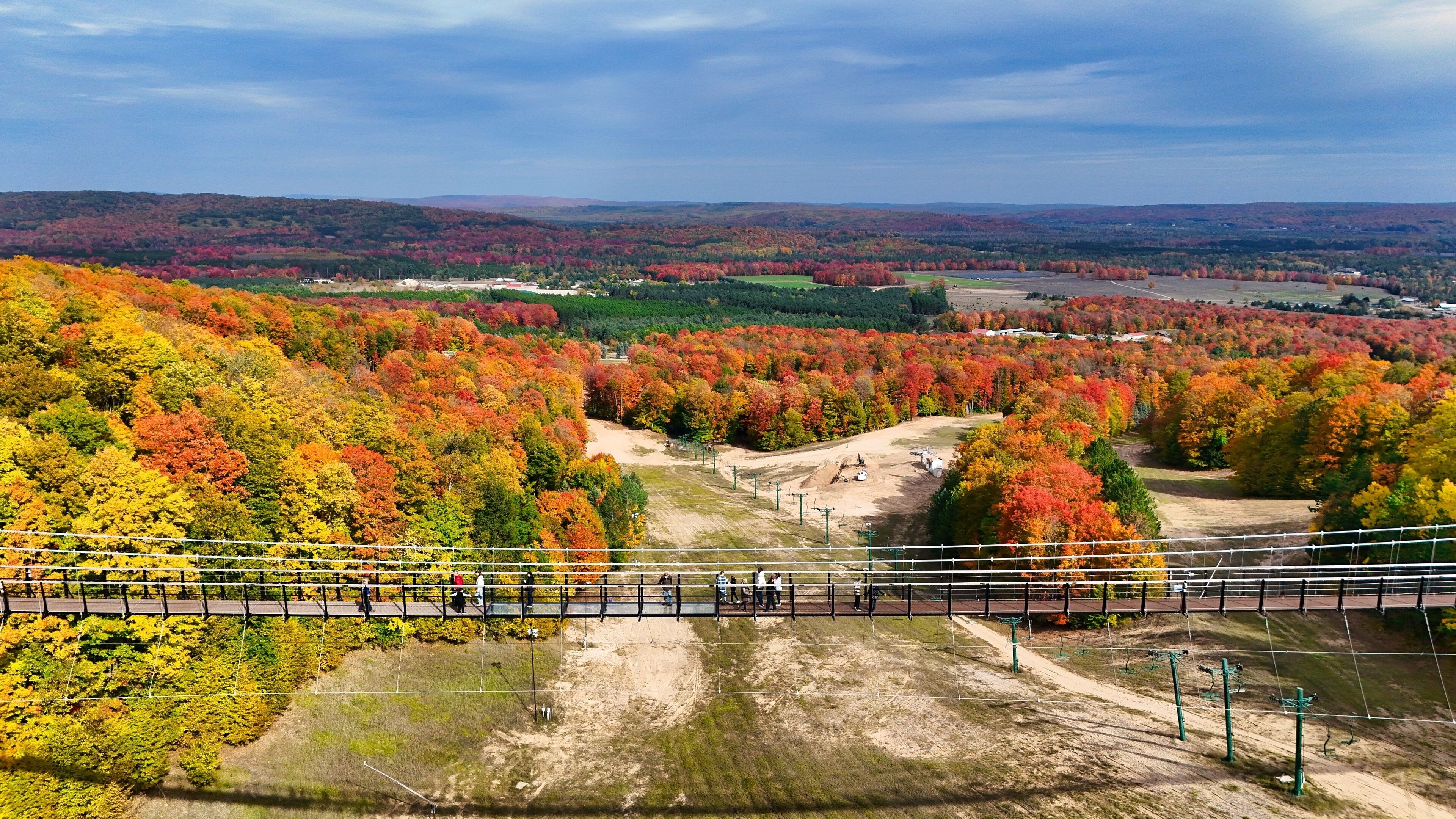A scenic view of a mountainous area with autumn foliage, a suspension bridge, and a distant body of water.