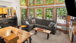 Ein modernes Wohnzimmer mit einer Eckcouch, einem Holztisch und einem Esstisch. Große Fenster sorgen für natürliches Licht und einen Blick ins Freie.