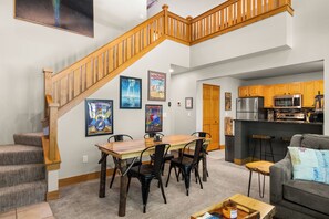 A spacious living area with a wooden staircase, a dining table with chairs, a kitchen with modern appliances, and artwork on the walls.