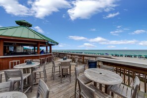 A wooden deck with round tables and chairs, a bar area, and a view of the beach and ocean. - Live the High Life: Luxe 11th Floor Gulf Front Condo Awaits Your Arrival! (Destin)