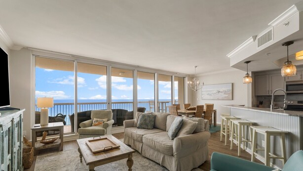 Amplia sala de estar con un ventanal que da al mar, una cocina con taburetes de bar y una zona de estar acogedora con un sillón color turquesa.