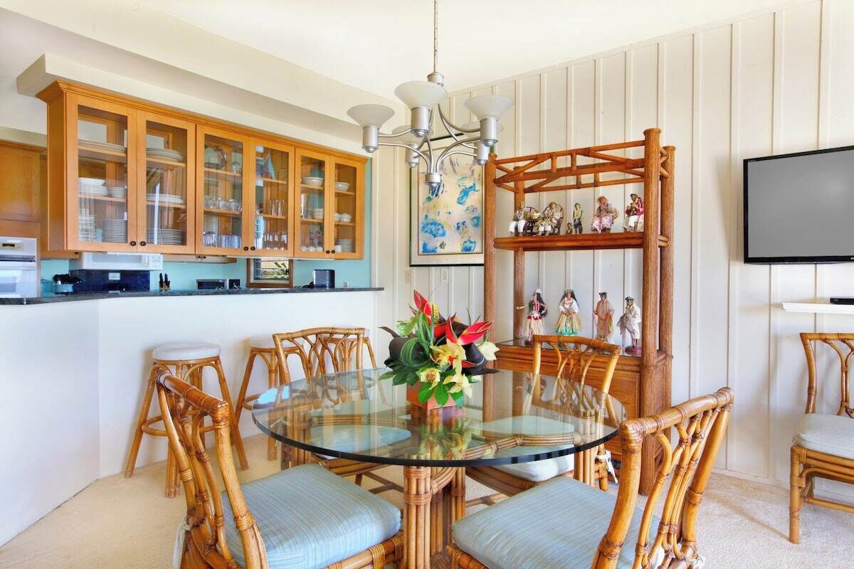 A dining area with a round glass table surrounded by wicker chairs, a wooden cabinet with glass doors, and a television mounted on the wall.
