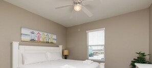 Un dormitorio con una cama, un ventilador de techo, un cuadro de casas de playa, una ventana con persianas y una planta.