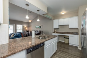 A modern kitchen with granite countertops, white cabinetry, and a dishwasher. There is a ceiling fan, pendant lights, and a sign on the wall.