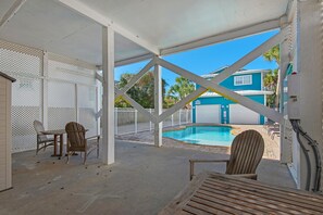 Un espacio al aire libre cubierto con una piscina, sillas de madera y vistas a un edificio azul.