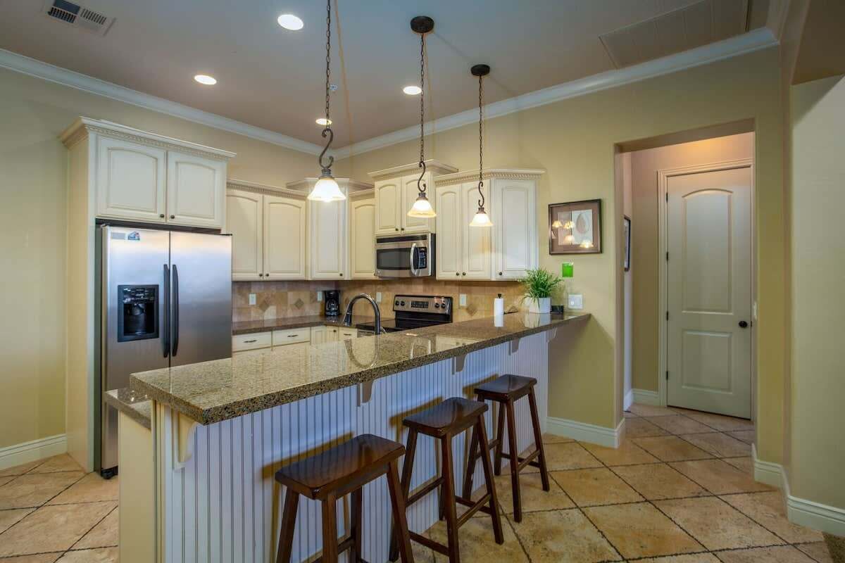 Una cocina con armarios blancos, un refrigerador de acero inoxidable, un microondas y una encimera de granito. Hay tres taburetes de madera en la isla.