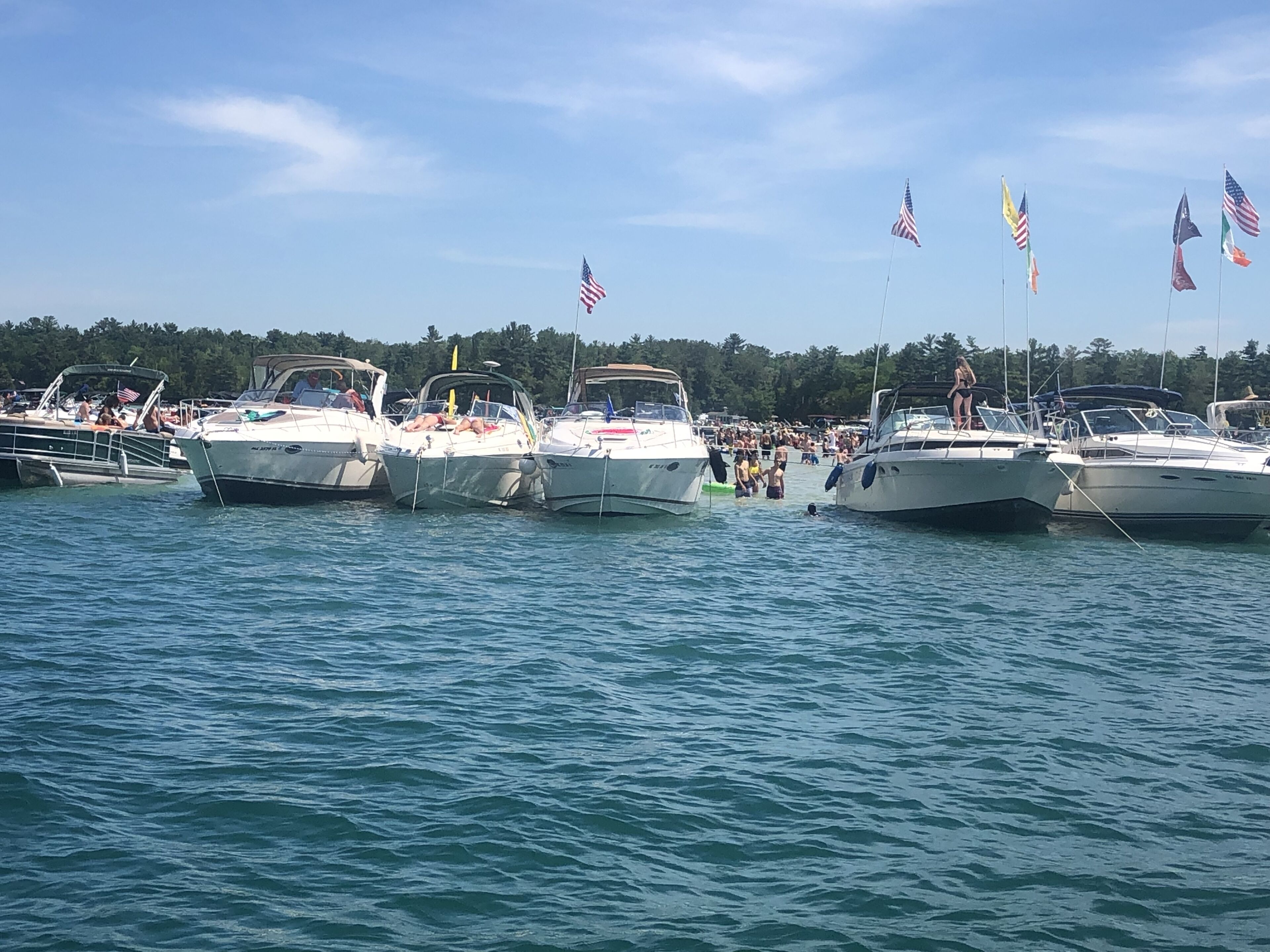 A marina with several boats docked, a crowd of people, and flags flying.