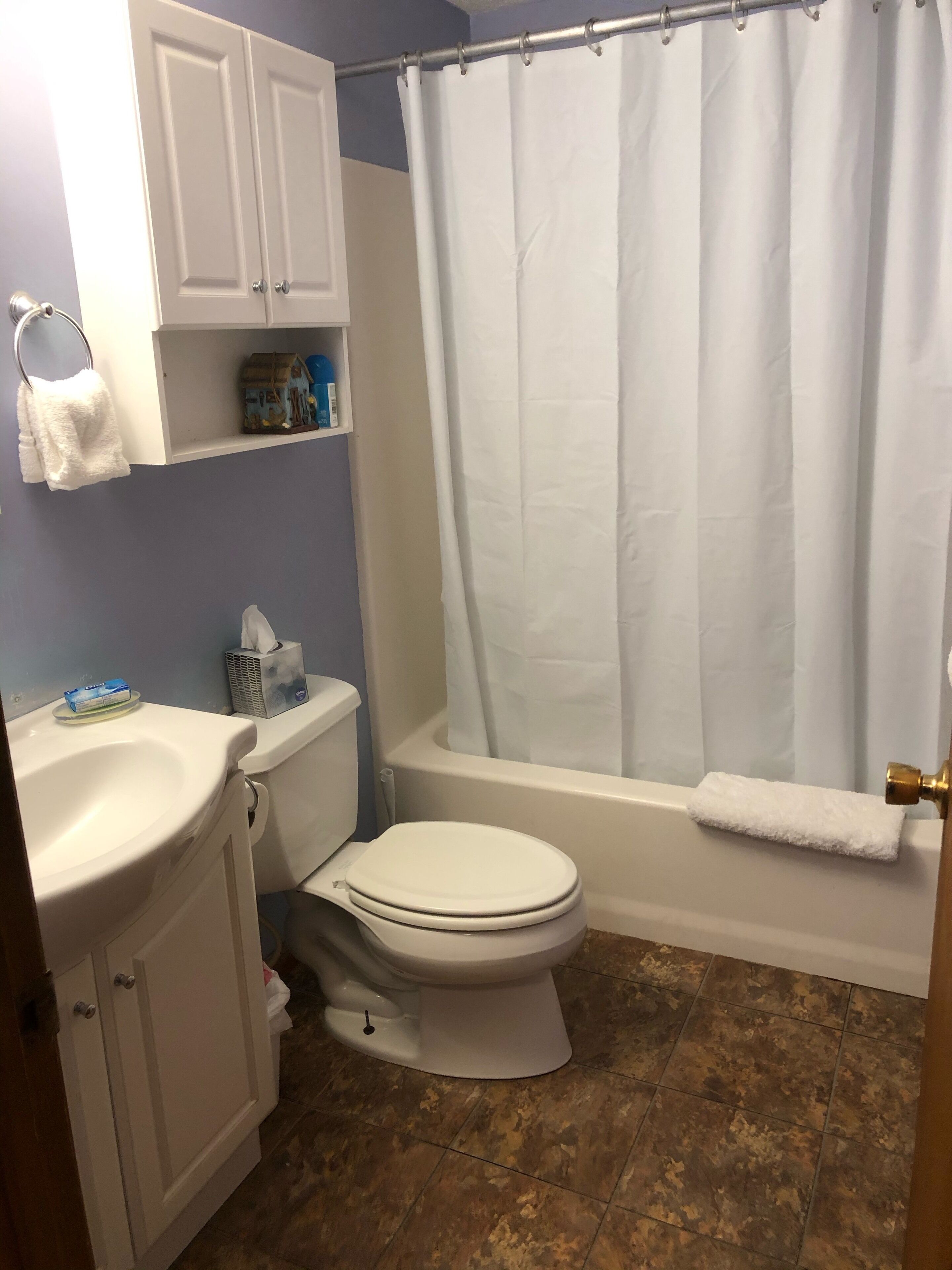 Un baño con inodoro, lavamanos y bañera. Hay un perchero sobre el lavamanos y una cortina de ducha en la bañera.