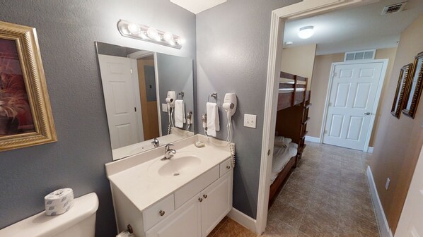 A bathroom with a sink, mirror, and hairdryer. There is a door leading to a bedroom with a bunk bed.