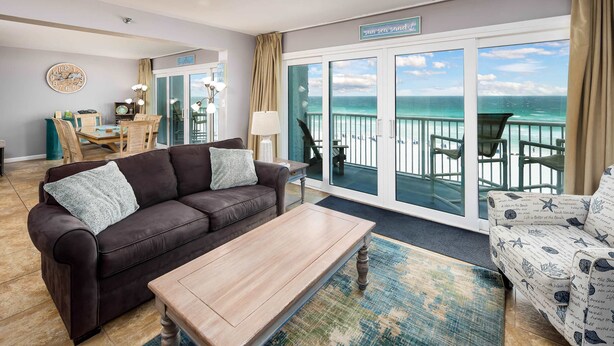 A living room with a brown leather sofa, a wooden coffee table, and a patterned rug. There is a dining table with chairs and a clock on the wall. A large sliding glass door leads to a balcony with a view of the ocean.