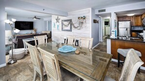 A dining area with a glass-top table, wooden chairs, and a kitchen in the background.