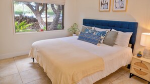 A neatly made bed with a blue headboard, a bedside table with a lamp, and a window with a view of a tree and a building. - Ma'alaea Surf Unit C1 Beachfront Condo, 2BR Ground Floor, AC, Wifi, Kitchen (Kihei)