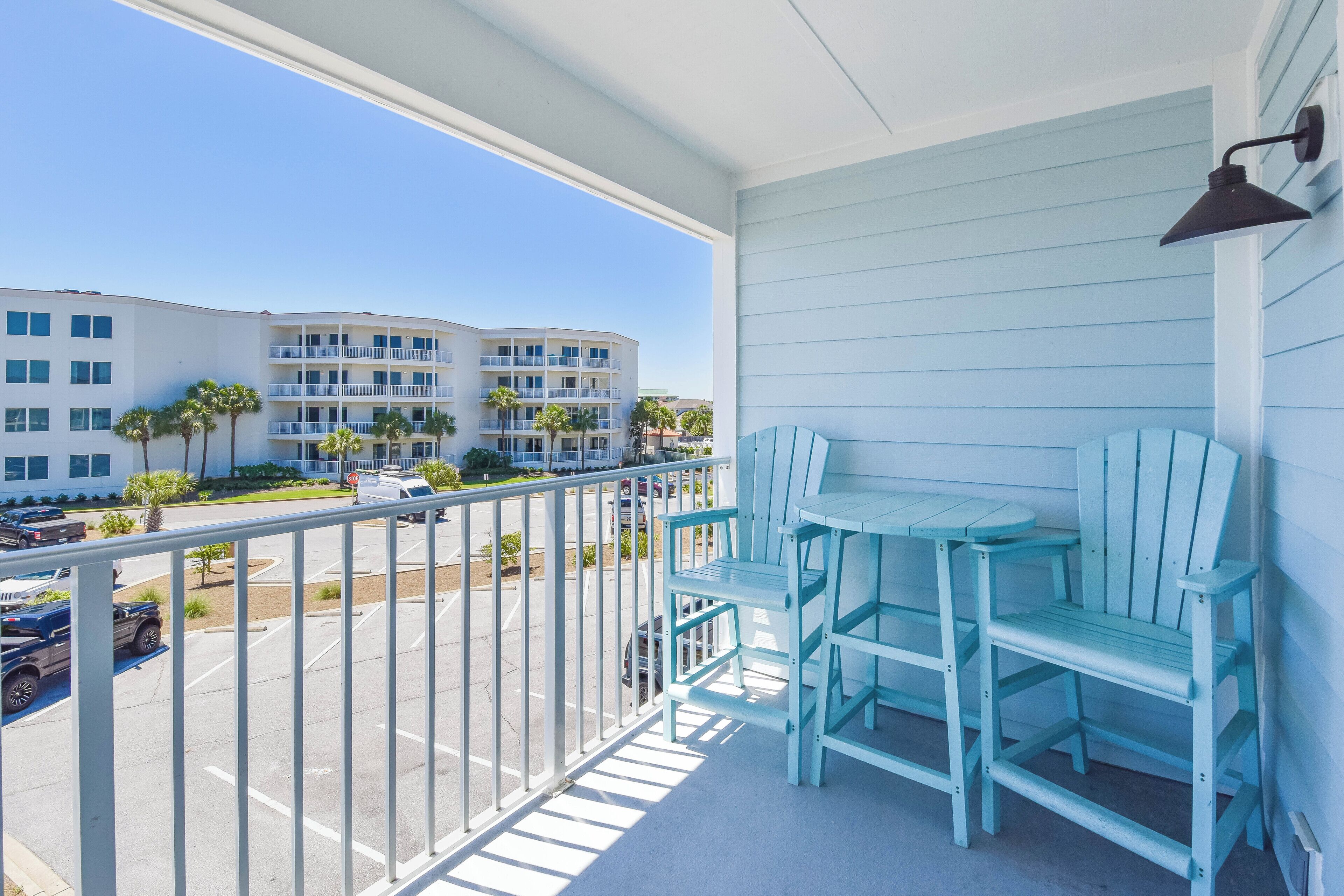 A balcony with a table and chairs, a view of a parking lot, and buildings in the background.