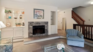 A living room with a fireplace, a blue armchair, and a coffee table with flowers. - Beachwalk 174 - Shipyard 3BR/3.5 BA Townhouse (Hilton Head Island)