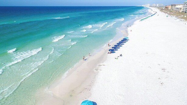 Une plage à l’eau turquoise et limpide, avec un rivage sablonneux et une rangée de parasols bleus.
