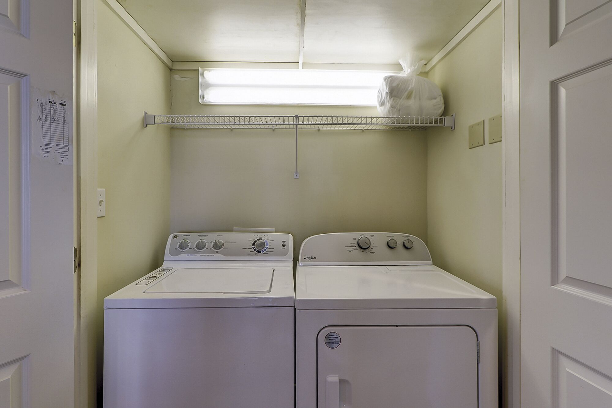 Un lavadero con dos lavadoras y secadoras Whirlpool, un estante metálico sobre las secadoras, y una pequeña ventana que proporciona luz natural.