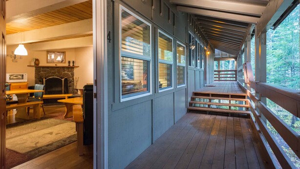 A wooden deck with a stone fireplace, a dining area with a table and chairs, and large windows offering a view of the outdoors. - Summer special, stay before 9/8/26 get third night free ! (Government Camp)