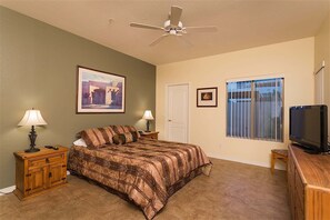 Ein Schlafzimmer mit einem Bett, zwei Nachttischen, einem Deckenventilator, einem Fernseher, einer Tür und einem Fenster mit Gitterstäben.