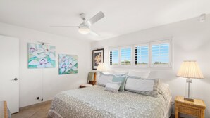 A bedroom with a bed, a ceiling fan, artwork on the walls, a window with blinds, and a bedside lamp.