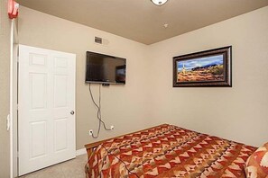 Un dormitorio con cama, televisor montado en la pared, puerta y un cuadro enmarcado.