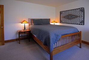 Un dormitorio con una cama de madera, dos mesitas de noche con lámparas, un adorno en la pared y piso alfombrado.