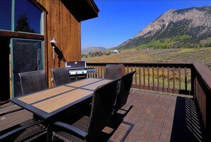 Una terraza de madera con una mesa, sillas, una parrilla y vistas a un paisaje montañoso.