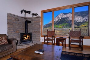 Una sala de estar acogedora con chimenea de piedra, dos sillones de madera, un sofá de cuero marrón y un amplio ventanal con vistas a la montaña.