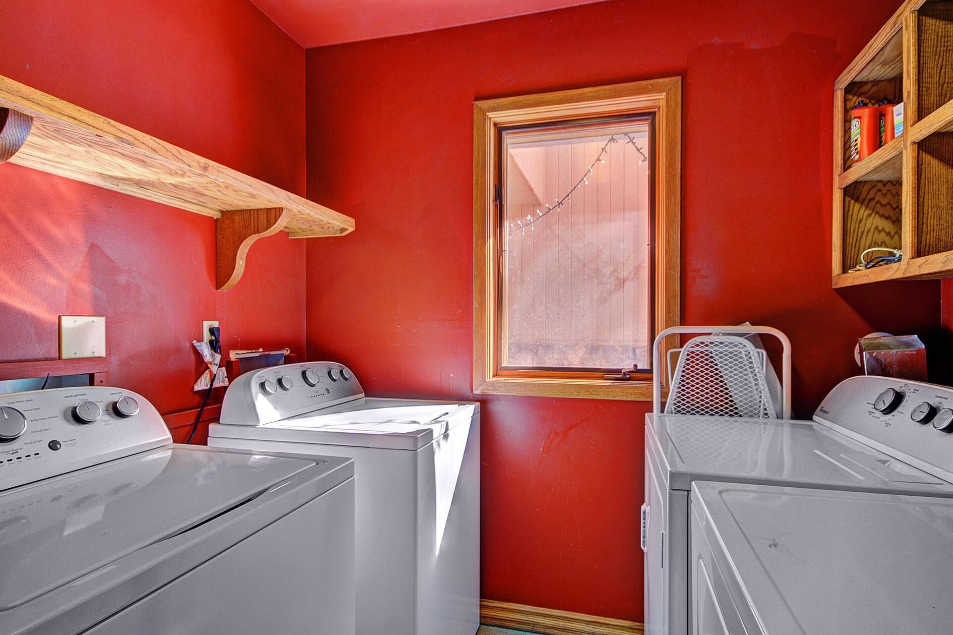 A laundry room with two white washers and dryers, a wooden shelf, a window with a curtain, and a metal rack.