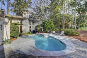 Piscina com borda curva, cercada por um terraço pavimentado. Há mobiliário de exterior e uma área de churrasco coberta.