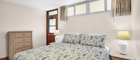 Un dormitorio con una cama grande, un ventilador de techo, una ventana con cortinas, una mesita de noche con lámpara y un aparador de madera.