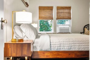 A bedroom with a wooden bed, a bedside table with a lamp, and two windows with blinds.