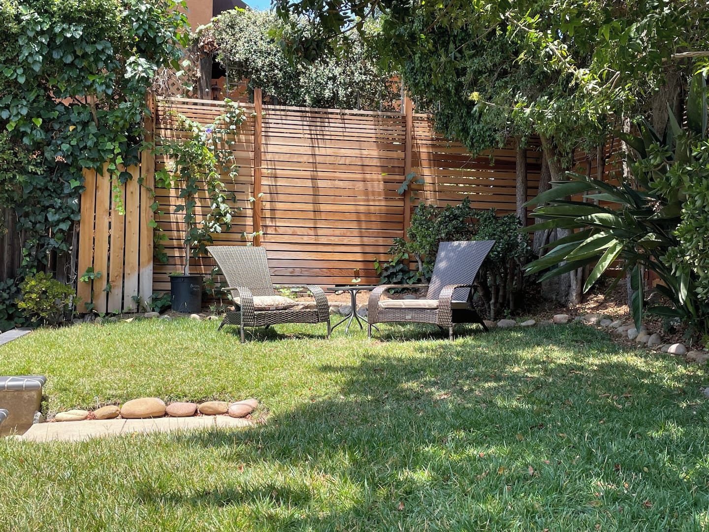 A backyard with a wooden fence, two wicker chairs, a small table, and a potted plant.