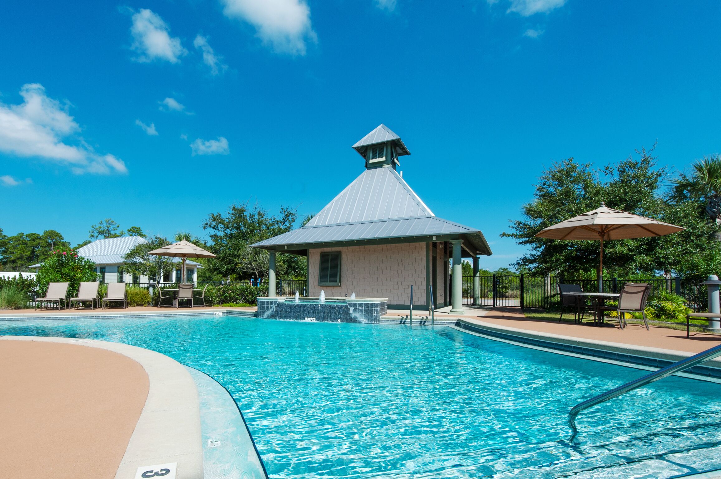 A swimming pool with lounge chairs and umbrellas, surrounded by greenery and a clear sky.