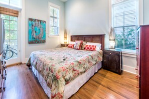 A bedroom with a large bed, wooden floor, a nightstand with a lamp, a dresser, and a window with blinds. - ☀️Close to Beach-Bikes-Community Pool-Surfin' Turtle (Santa Rosa Beach)