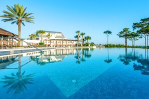A clear blue infinity pool with lounge chairs and palm trees, reflecting a modern resort building. - ☀️Close to Beach-Bikes-Community Pool-Surfin' Turtle (Santa Rosa Beach)