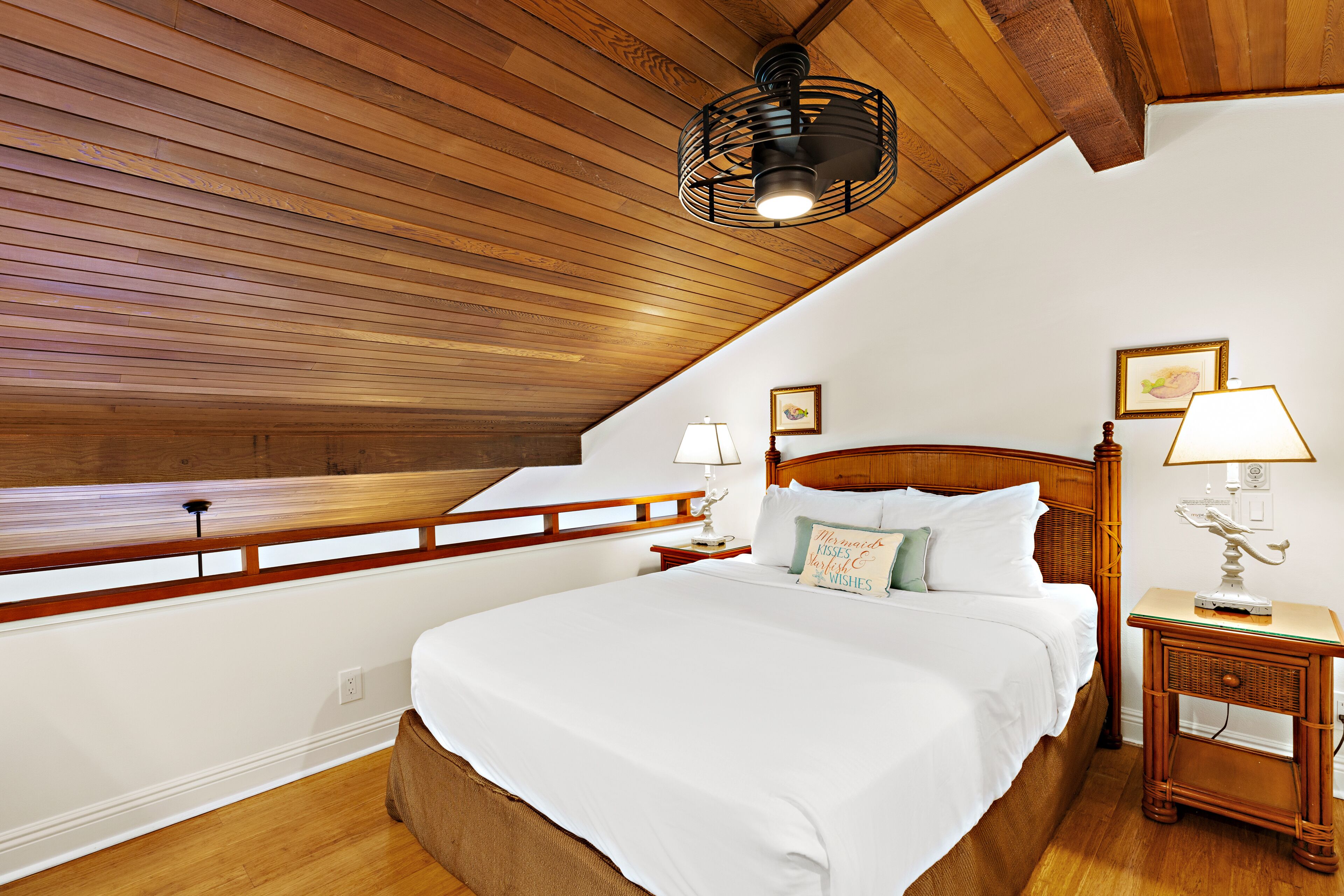 Une chambre avec un plafond en pente, des poutres en bois, un grand lit avec du linge de lit blanc, deux tables de chevet avec des lampes et une petite étagère fixée au mur.