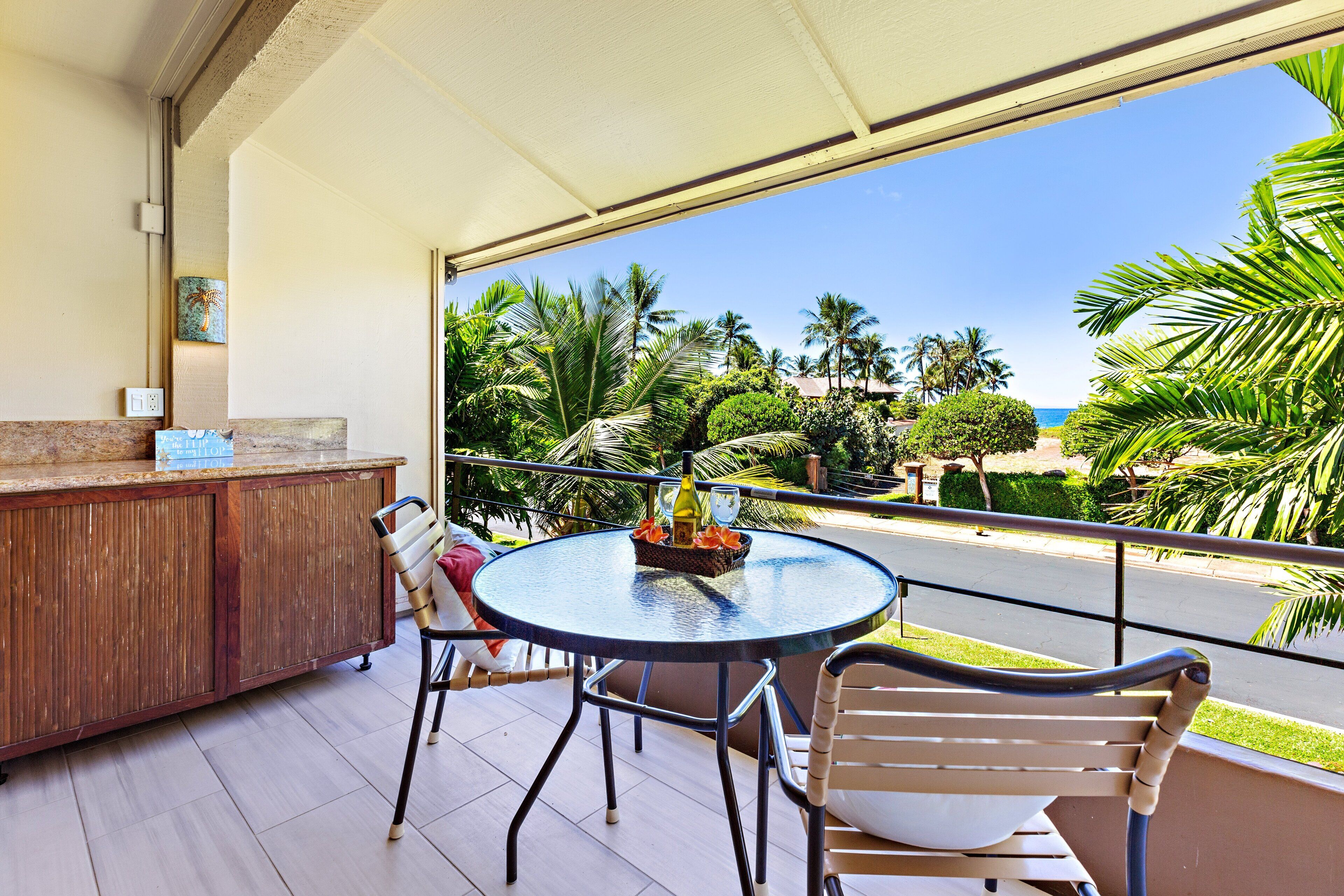 A balcony with a round table, chairs, a wooden cabinet, and a view of palm trees and the ocean.