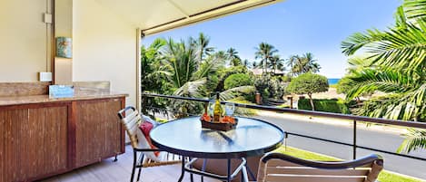 A balcony with a round table, chairs, a wooden cabinet, and a view of palm trees and the ocean.