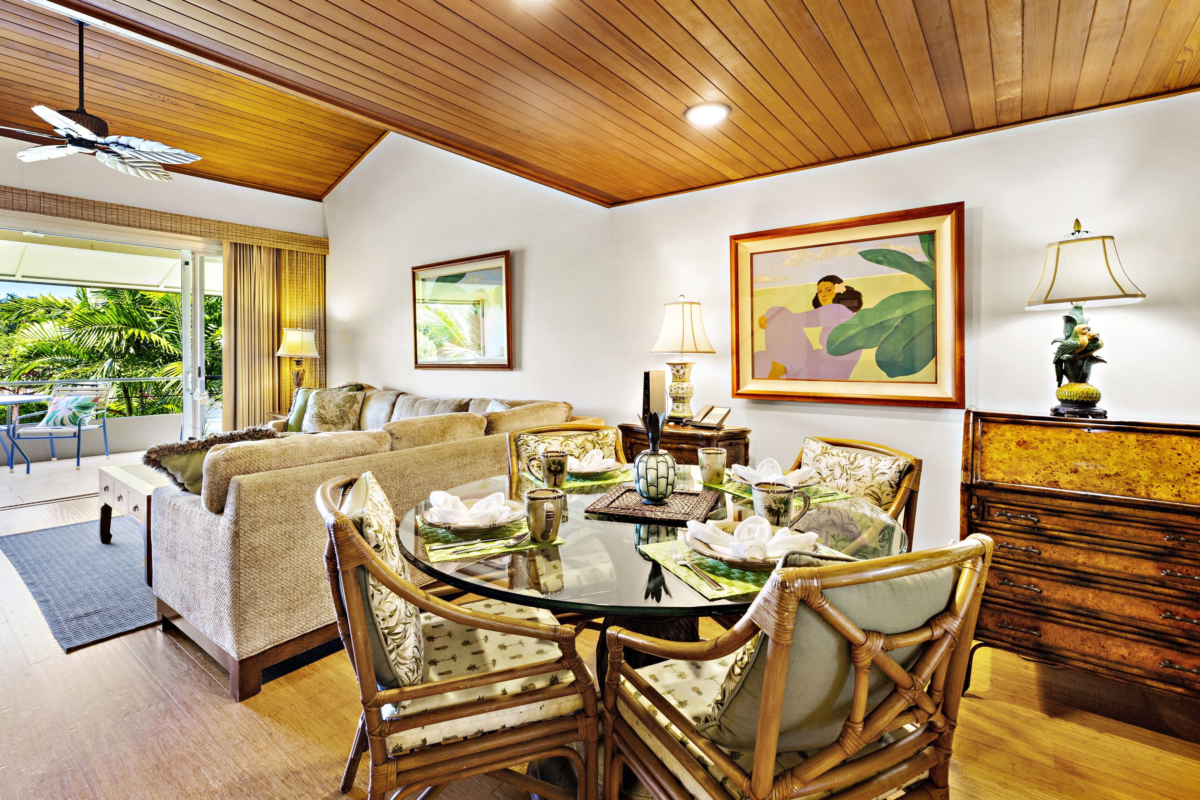 A well-lit room with a dining table set for four, surrounded by wicker chairs. There is a sofa and a small table with a lamp in the background.