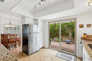 Une cuisine équipée d’un réfrigérateur en acier inoxydable, de meubles blancs, d’un coin repas avec des chaises en bois et d’une porte-fenêtre coulissante donnant sur une terrasse avec vue sur la verdure.