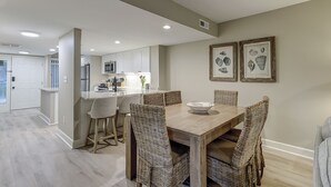Zona pranzo con tavolo in legno e sedie in vimini, cucina sullo sfondo e quadri appesi al muro.