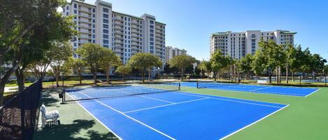 An outdoor tennis court with a net, surrounded by trees and buildings.